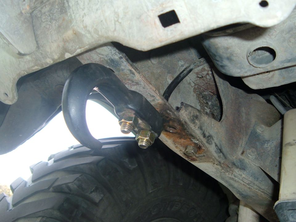Close-up of a black tow hook bolted to a rusty metal bracket on a vehicle's undercarriage, with a tire visible below