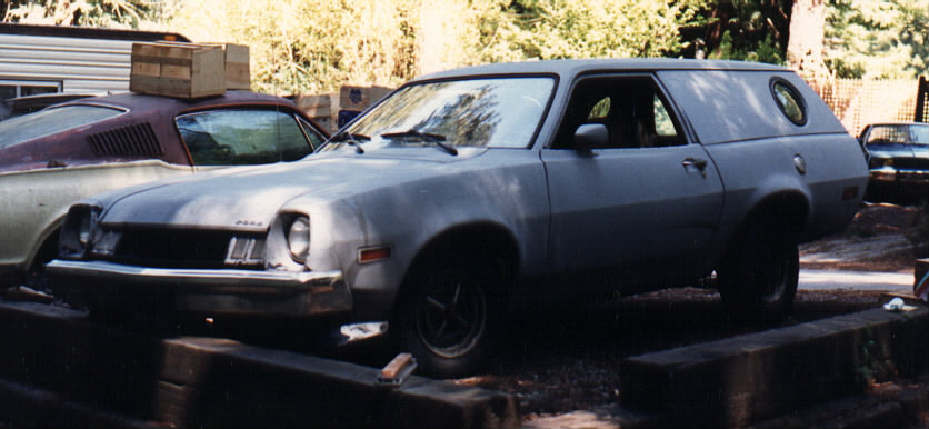 Gray primered Ford Pinto wagon with porthole rear window parked in a wooded dirt yard