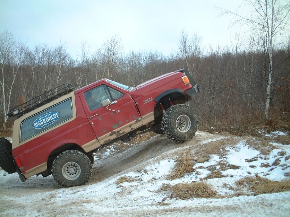 Red and tan lifted Ford Bronco climbing a snowy dirt mound in profile view