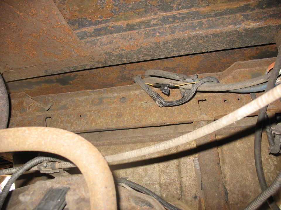 Underbody of a rusty vehicle showing frame rails, exhaust pipe, and wiring harness
