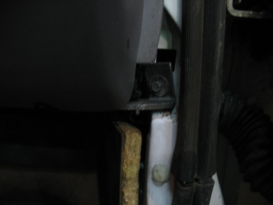 Close-up of a metal bracket with a bolt, attached to a dark grey panel and white trim inside a vehicle