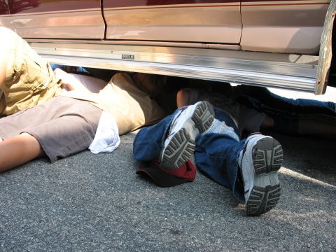 1995 Ford C-350 (Centurion Conversions) - People are working or resting underneath a vehicle, with their legs and feet visible on the asphalt.