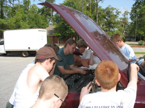 1995 Ford C-350 (Centurion Conversions) - A group of young men are gathered around the engine bay of a car with its hood open, seemingly working on it.