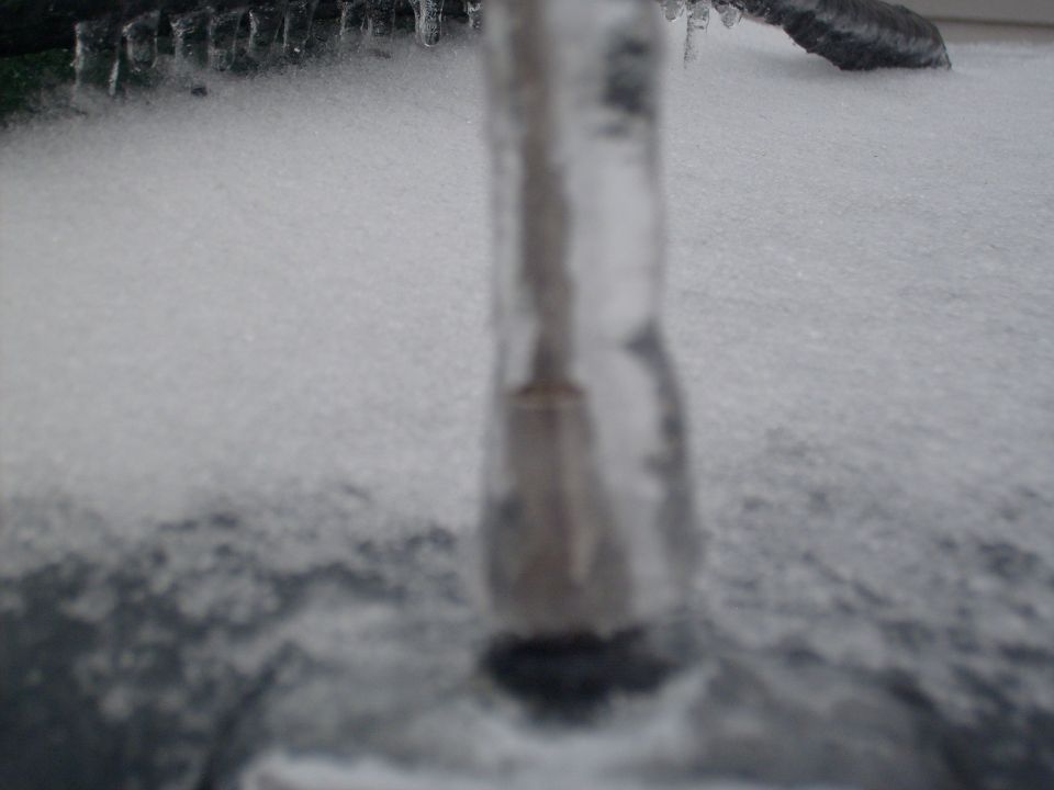 Close-up of an icicle hanging over snow with a dark, blurred object in the background