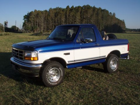 1996 Ford Bronco - A two-tone blue and white 1996 Ford Bronco sits in a grassy field under a clear blue sky.