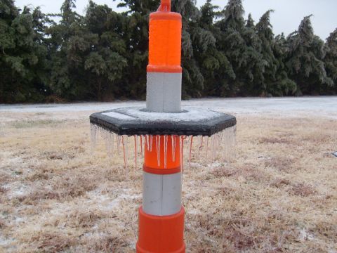 1995 Ford C-350 (Centurion Conversions) - A traffic cone stands covered in ice and icicles during an apparent ice storm, with frozen trees in the background.