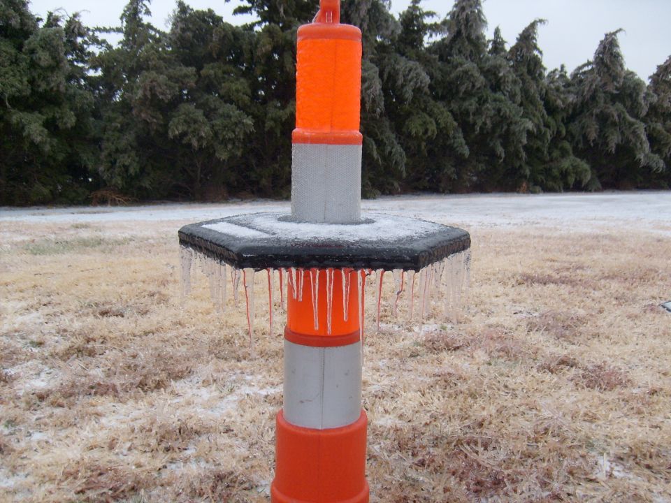 Orange and gray traffic cone with icicles hanging from its base, surrounded by frozen grass and trees