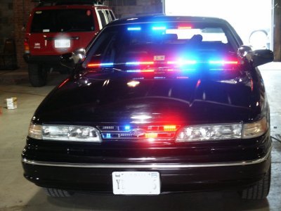 2001 Ford Crown Victoria - A black 2001 Ford Crown Victoria police car is shown with its emergency lights activated, with a red SUV in the background.
