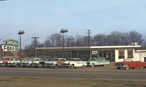 1968 Shelby GT500 - Vintage Ford dealership lot displays multiple 1950s vehicles under a bright sky.