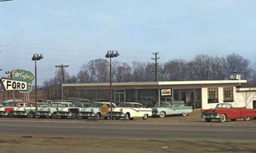 Ford dealership lot with several 1950s cars, including a red convertible and a light blue sedan