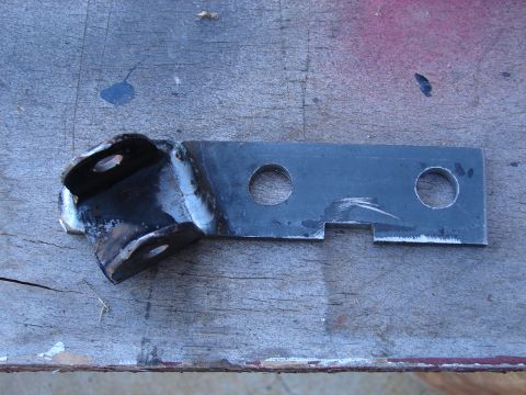 1973 Ford Ranchero - Fabricated steel bracket lying on a plywood workbench: a flat bar with two drilled holes and a notched end, freshly welded to a black forme…