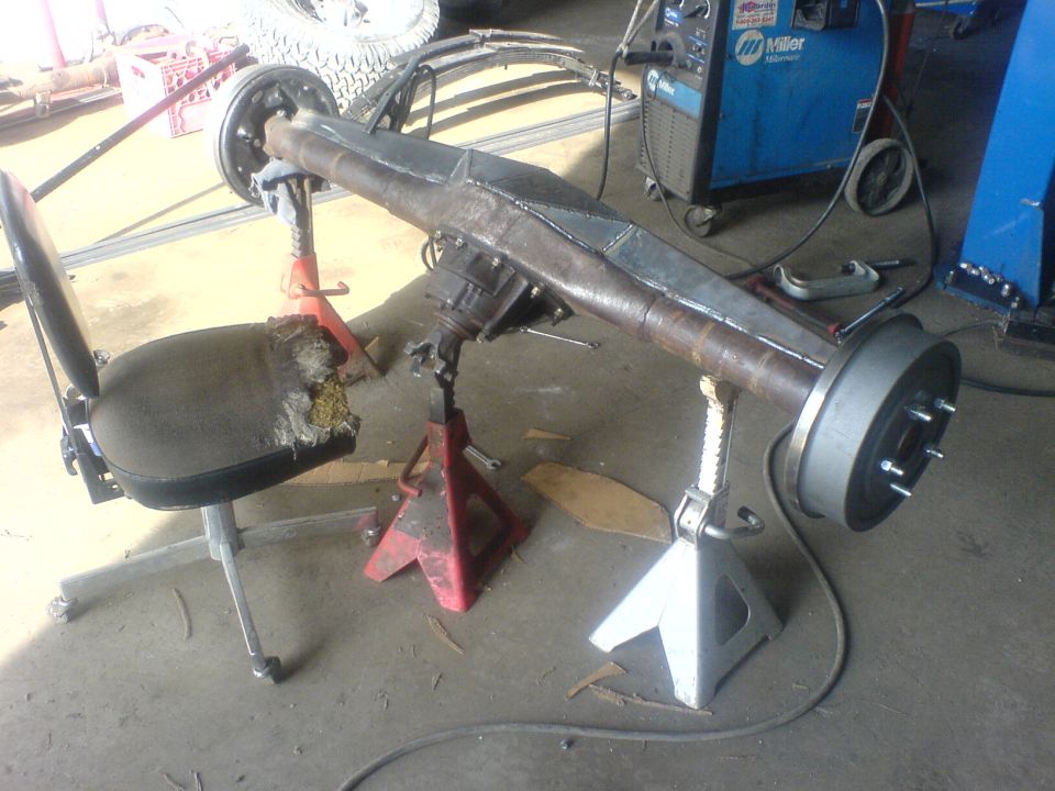 Rear axle assembly on jack stands with a torn office chair nearby and a welding machine in the background