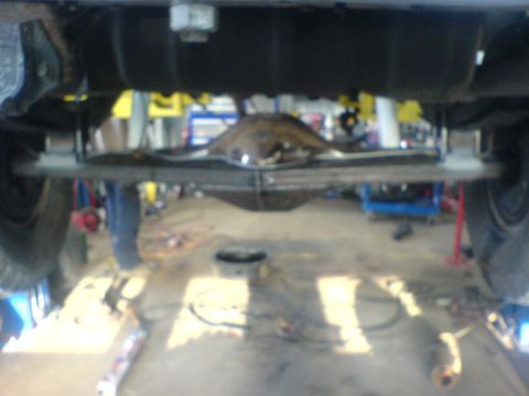 1985 Ford Bronco - The underside of a vehicle is shown, focusing on the rear axle and suspension system in a workshop setting.