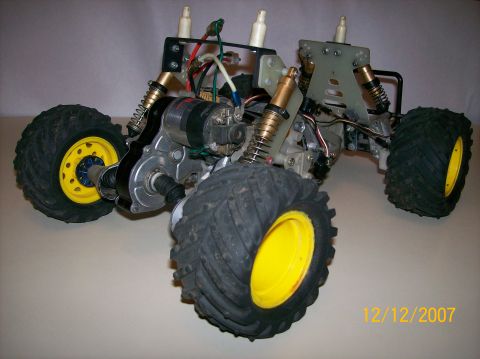1979 Ford Bronco - A stripped RC monster truck chassis viewed low from the front, with gold-anodized coil-over shocks, a silver alloy gearbox and electric mot…