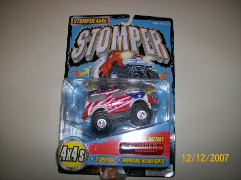1979 Ford Bronco - A sealed Stomper 4x4s battery-powered toy truck on its blister card, holding a lifted SUV-bodied truck painted in a red, white and blue Ame…