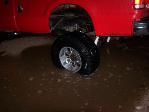1991 Ford Bronco - A red truck's rear wheel is submerged in muddy water, with the chrome rim partially visible.