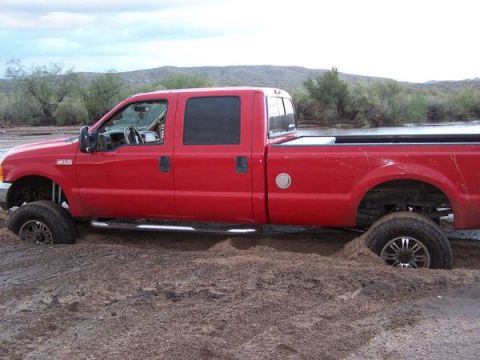 1991 Ford Bronco - Red Ford F-250 Super Duty pickup truck is stuck in mud, showing its lifted suspension and aftermarket wheels.