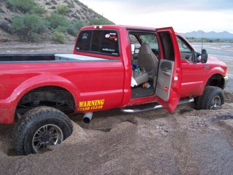 1991 Ford Bronco - A red pickup truck is stuck in soft sand with its doors open, showing the interior and the wheels buried.