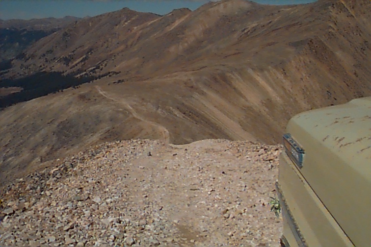 Mountain landscape with a dirt road and the corner of a tan vehicle visible on the right. Watermark reads SUPERMOTORS.NET