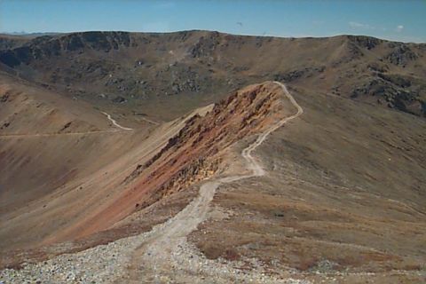 1994 Ford Bronco - A remote mountain pass with a dirt road snaking along the ridge, under a vast expanse of blue sky.