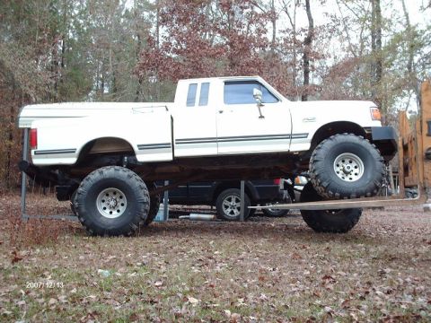 1990 Ford F-150 - This heavily modified pickup truck features oversized tires and a significant lift kit, with another vehicle partially visible beneath it.