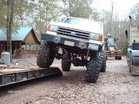 1990 Ford F-150 - A heavily modified 1990 Ford F-150 pickup truck with oversized tires is shown in a 3/4 front view, with other vehicles and a building in th…