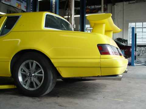 1986 Mercury Cougar - This modified yellow coupe features a prominent rear spoiler and dual exhaust tips, with a Supermotors logo visible.