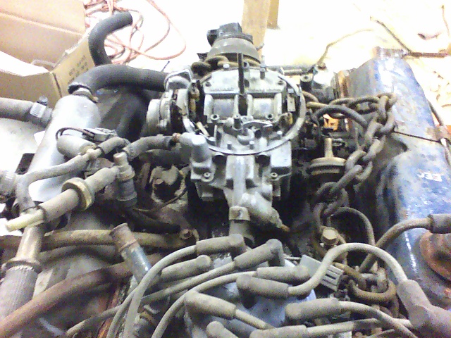 Close-up of an engine block with a carburetor, spark plug wires, and hoses. A chain is wrapped around part of the engine