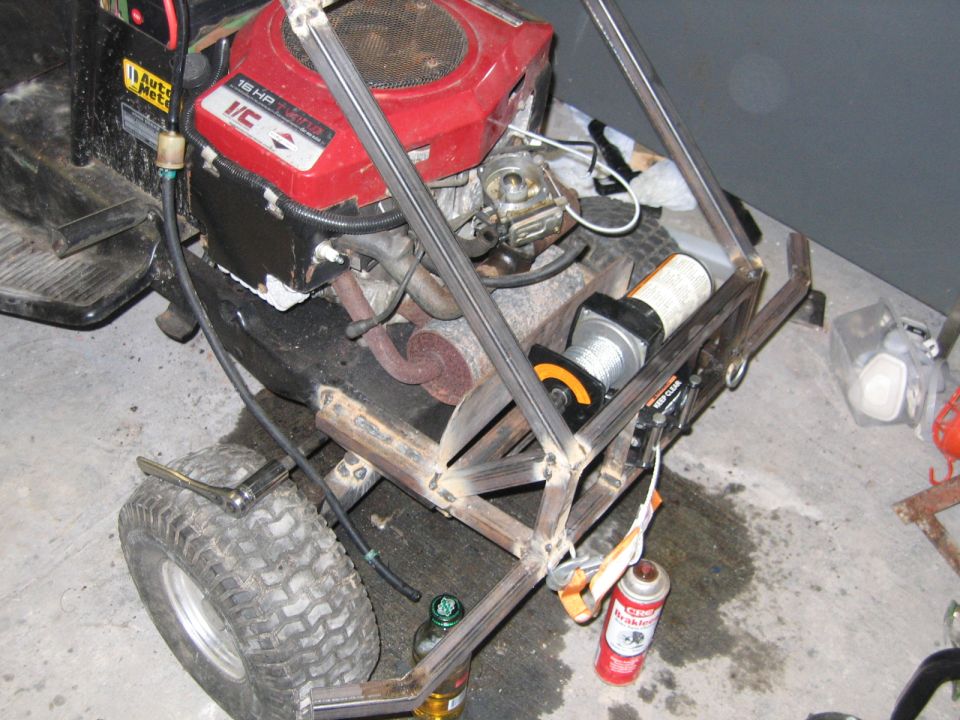 Modified lawnmower with a winch and custom metal frame. Red engine, black chassis, and large rear tires are visible