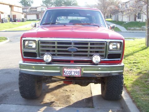 1986 Ford Bronco - The front grille and headlights of a red Ford Bronco are shown, with a California license plate mounted on the bumper.