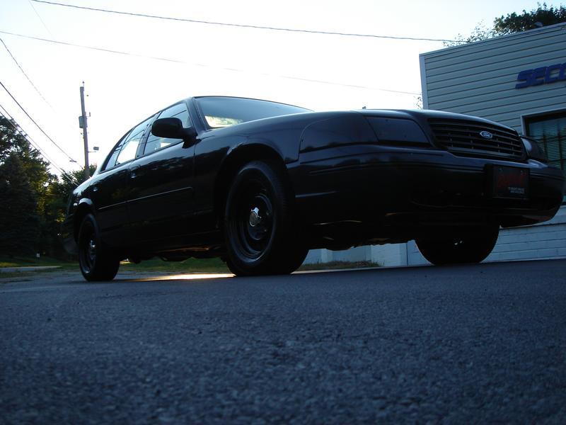 Low-angle view of a black Ford Crown Victoria sedan with black steel wheels parked on pavement