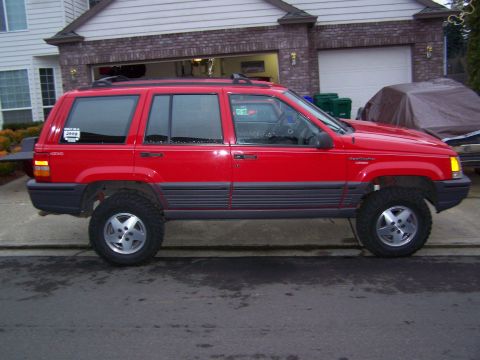 1994 Jeep Grand Cherokee - A red 1994 Jeep Grand Cherokee is parked in a driveway, showing its side profile with aftermarket wheels.