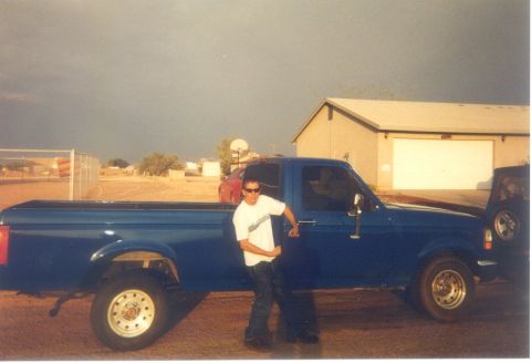 1992 Ford Bronco - Scanned snapshot of a blue 1990s Ford full-size regular-cab long-bed pickup shown in side profile on a dirt driveway, with a person in a wh…