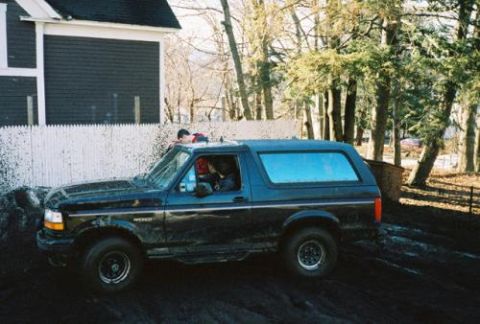 1995 Ford Bronco - A dark-colored Ford Bronco is seen in a muddy outdoor setting with trees and a house in the background.