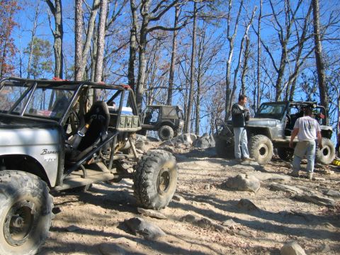 1992 Ford Mustang - Multiple off-road vehicles are parked on a rocky, wooded trail under a clear blue sky.