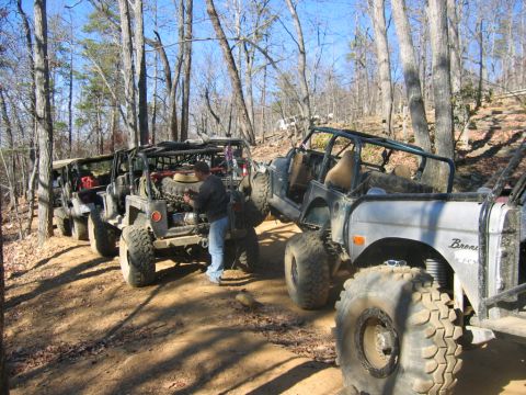 1992 Ford Mustang - Several modified off-road vehicles are parked on a dirt trail in a wooded area, with one vehicle's engine exposed.