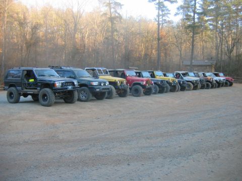 1992 Ford Mustang - Multiple off-road SUVs are parked in a line on a dirt lot with a wooded area behind them.
