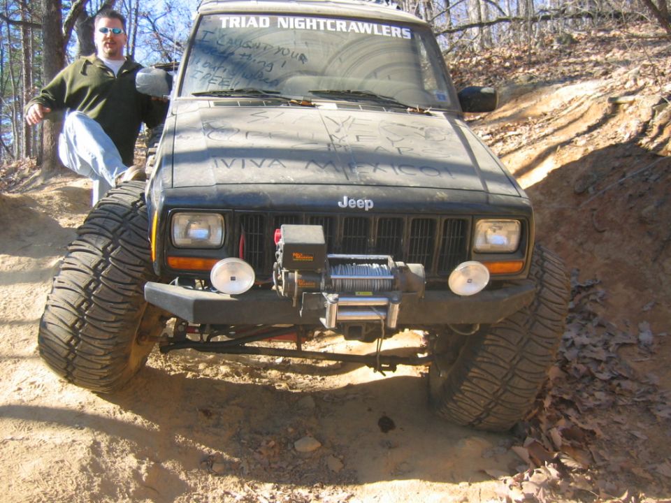 Man leaning on a lifted Jeep Cherokee XJ with a winch on the front, on a dirt trail