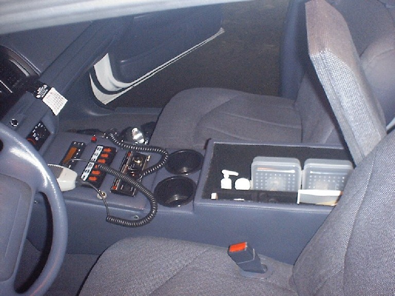 Interior shot of a police car's center console with radio equipment, cup holders, and storage. Includes a steering wheel on the left