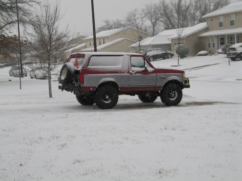 1995 Ford Bronco - Passenger-side view of a lifted red-and-silver two-tone Ford Bronco parked on a snow-covered suburban street during snowfall, snow dusting…