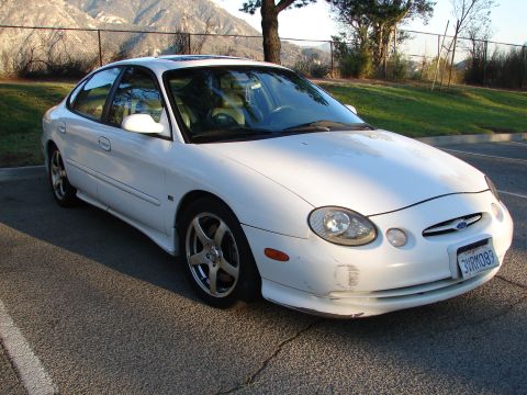 1993 Ford Taurus - White sedan with aftermarket wheels parked outdoors. Mountains and trees are visible in the background.
