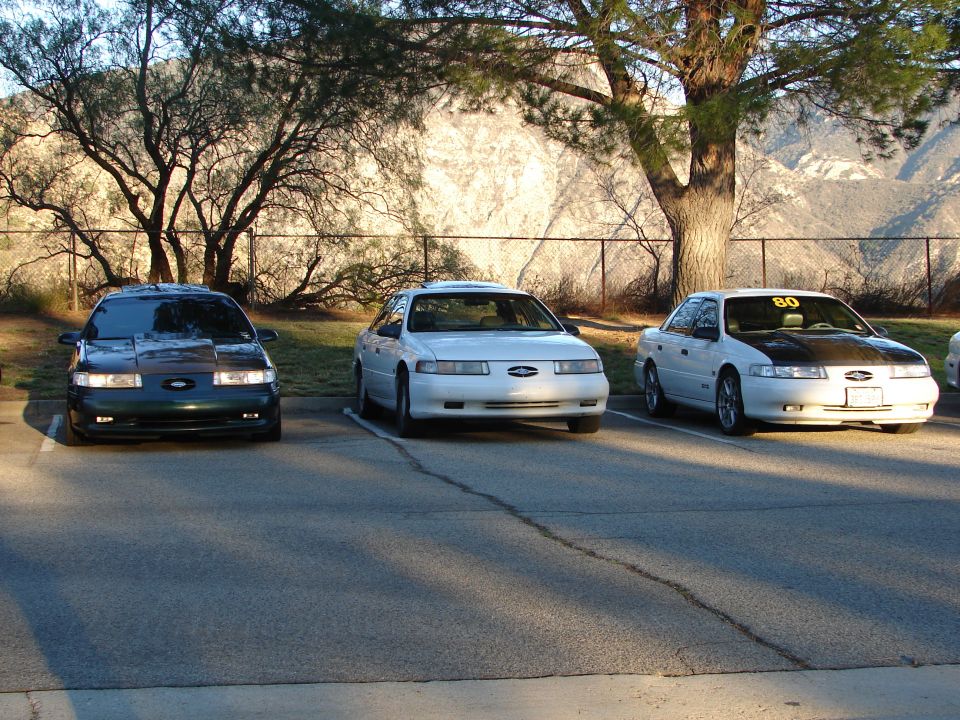 Three Ford Taurus sedans parked in a lot. The middle and right cars appear to be from the mid-1990s. The left car is darker and has a modified hood