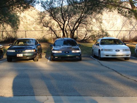 1993 Ford Taurus - Three Ford Taurus sedans are parked in a lot, with a hillside and trees in the background.