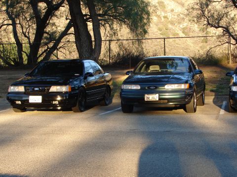 1993 Ford Taurus - Two Ford Taurus sedans are parked in a lot, one black and one green, with a hillside in the background.
