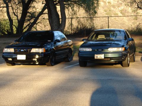 1993 Ford Taurus - Two Ford Taurus sedans are parked side-by-side in a lot, with trees and a fence in the background.