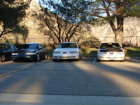 1993 Ford Taurus - Three sedans are parked in a lot with a mountainous backdrop and trees.