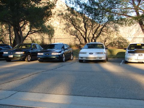 1993 Ford Taurus - Four sedans, including a 1993 Ford Taurus, are parked in a lot with trees and a rocky hillside in the background.