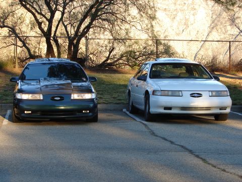 1993 Ford Taurus - Two Ford Taurus sedans are parked in a lot. The dark green Taurus has a modified hood.