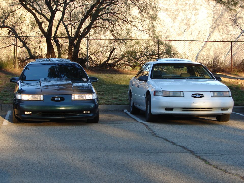 Two Ford Taurus sedans, one dark green and one white, parked in a lot. The dark green one has a modified hood