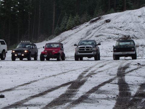1989 Ford Bronco - Several off-road vehicles are parked on a snow-covered ground, with a wooded hillside behind them.
